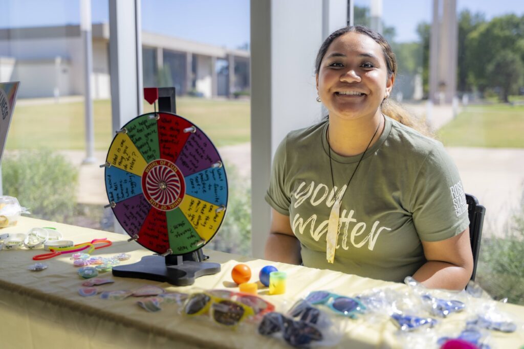 Smiling Rose State College staff member seated at a wellness activity table with a colorful prize wheel and giveaway items.