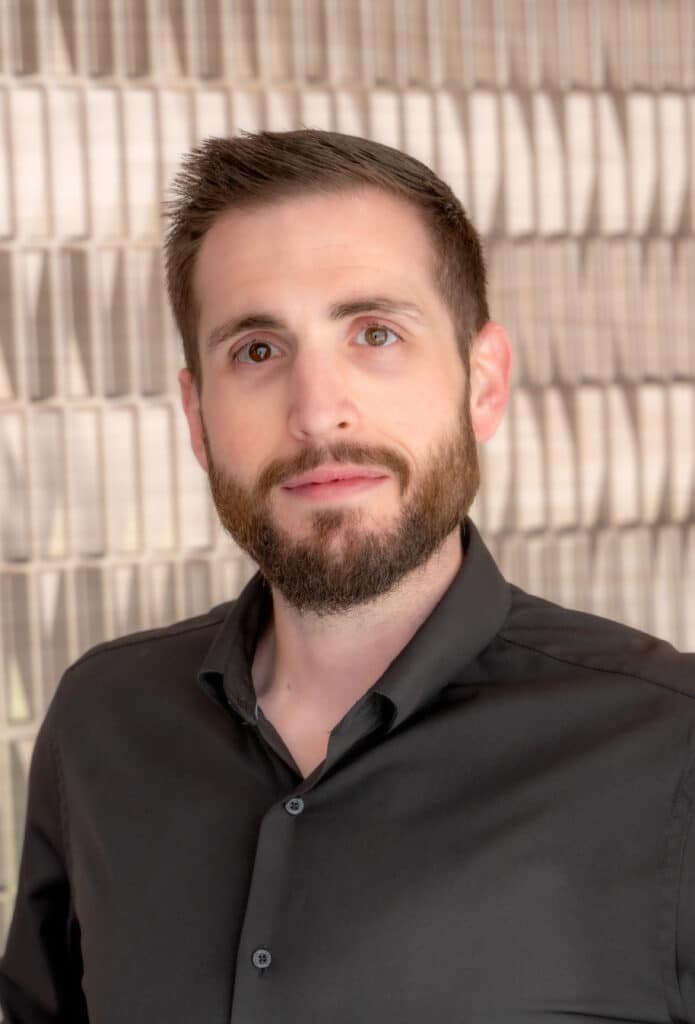 Professional headshot of a man with short brown hair and a trimmed beard wearing a black button-up shirt, in front of a softly blurred neutral background.