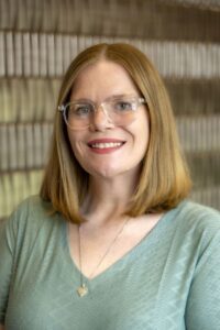 Portrait of a smiling person with shoulder-length light brown hair and clear-framed glasses, wearing a light green top and necklace, posed in front of a softly blurred patterned background.