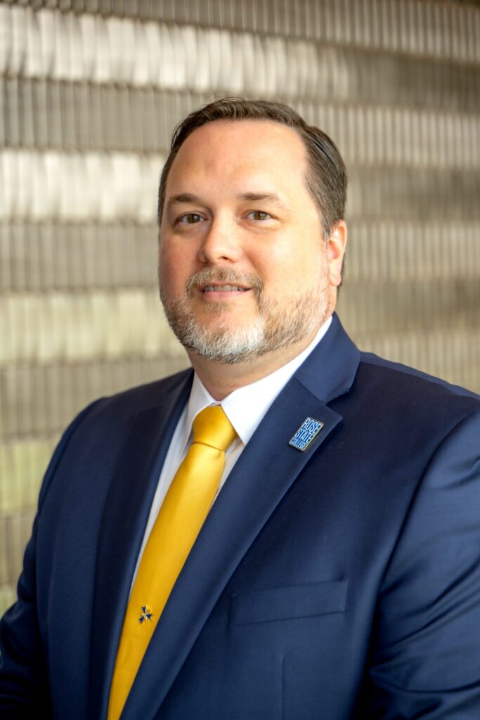 Professional headshot of Rose State College President Travis Hurst wearing a navy suit, white shirt, and gold tie, standing in front of a softly blurred architectural background.