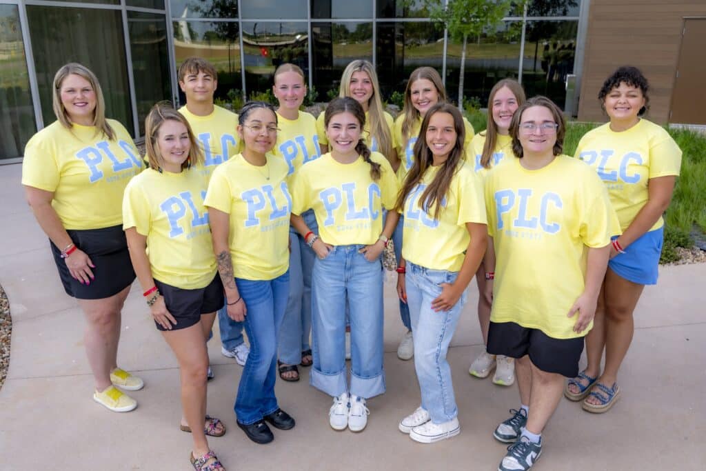 A group of RSC PLC students dressed in matching yellow shirts smile for a picture while standing outside of a building with glass windows.