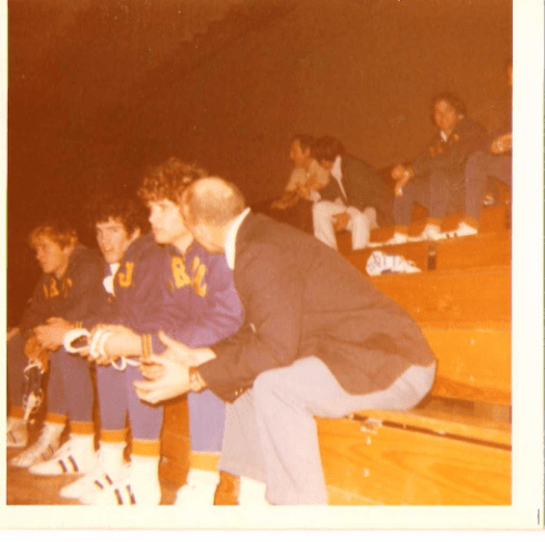 Vintage color photo of a wrestling coach speaking with players seated on wooden gym bleachers during a game, with other team members visible in the background.