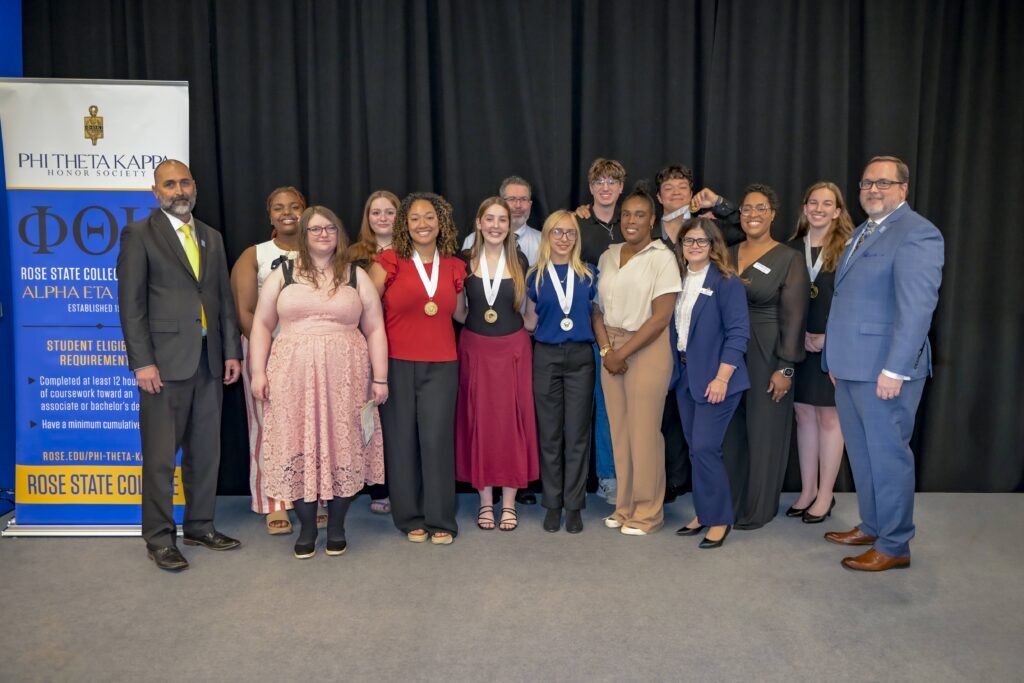 Group of students and faculty pose on stage at a Phi Theta Kappa induction ceremony, with several students wearing honor medallions and a banner for the Rose State College chapter displayed beside them.