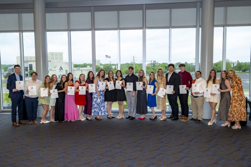 Group of students stand in a line indoors holding certificates and smiling, with large windows behind them overlooking campus.
