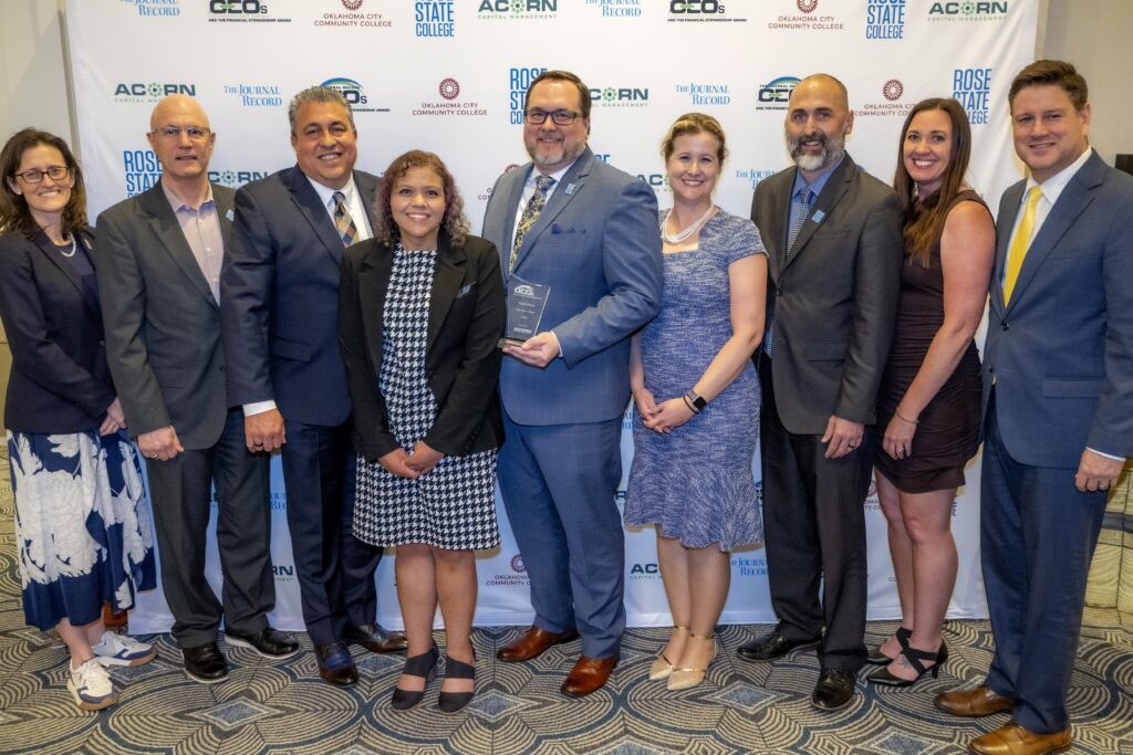 Group of nine professionals stand together in front of an event backdrop with partner logos, with one person holding an award plaque at a recognition ceremony.
