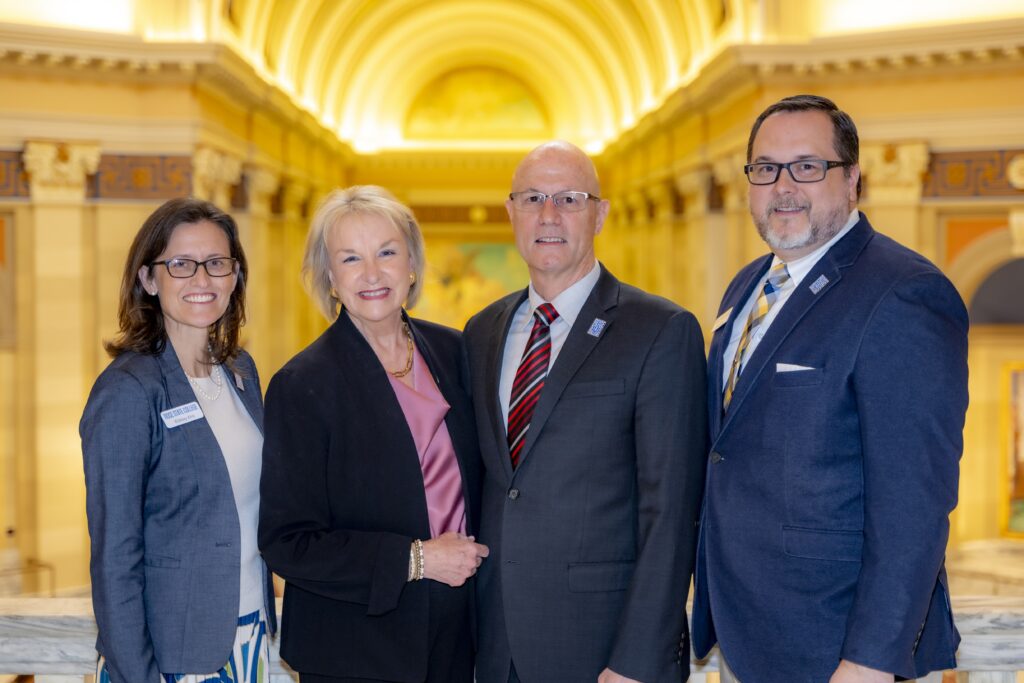 Four professionally dressed adults stand together smiling in an ornate, well-lit interior with arched ceilings and columns in the background.