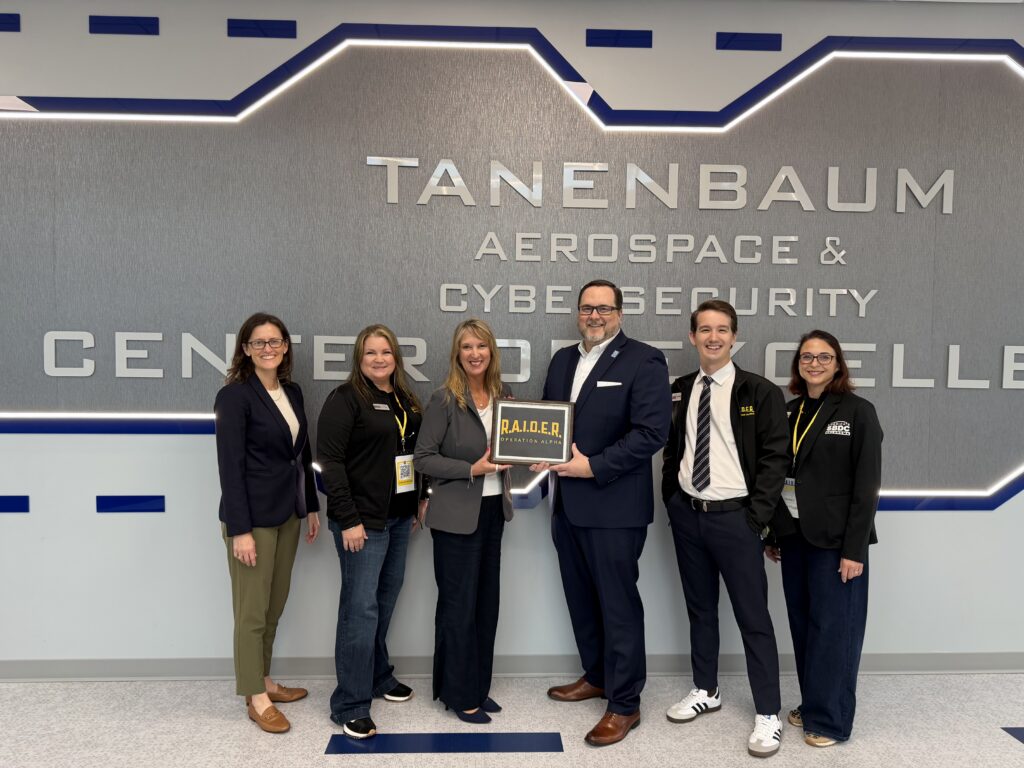 Group of faculty, staff, and a student stand inside the Tannenbaum Aerospace & Cybersecurity Center holding an “R.A.I.D.E.R.” award plaque and smiling for a photo.