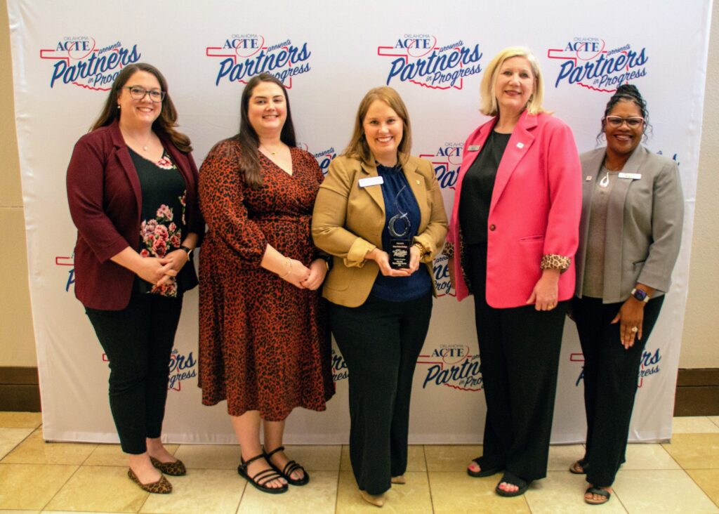 Five women stand together smiling in front of an Oklahoma ACTE “Partners in Progress” backdrop, with one woman holding an award.