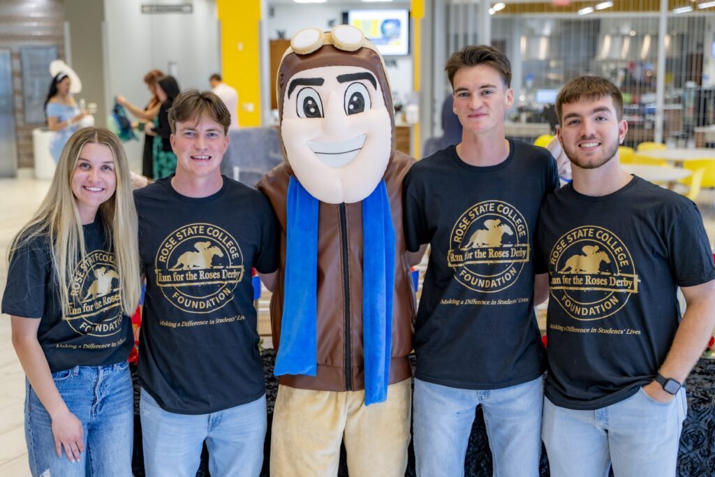 Four students wearing matching Rose State College Foundation “Run for the Roses Derby” T-shirts pose with the college mascot inside a campus building during a fundraising event.