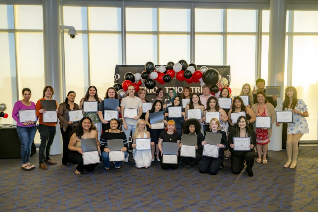 Large group of students pose indoors holding certificates in front of a black, red, and white balloon display at a TRIO awards banquet.