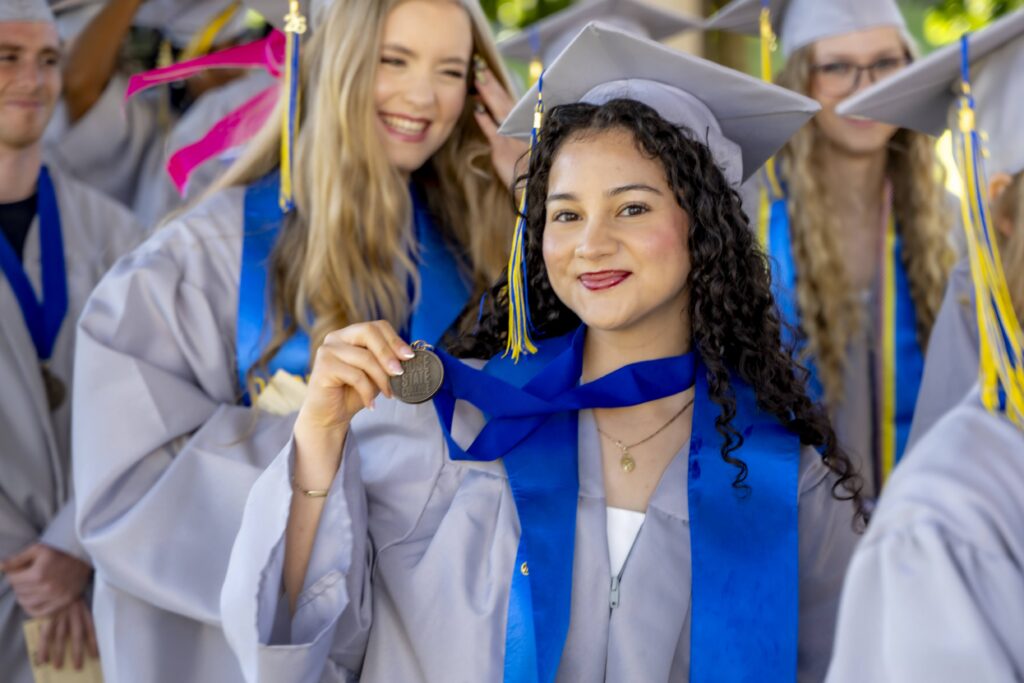 Rose State College graduate in cap and gown holds up a graduation medal while posing with fellow graduates during commencement.