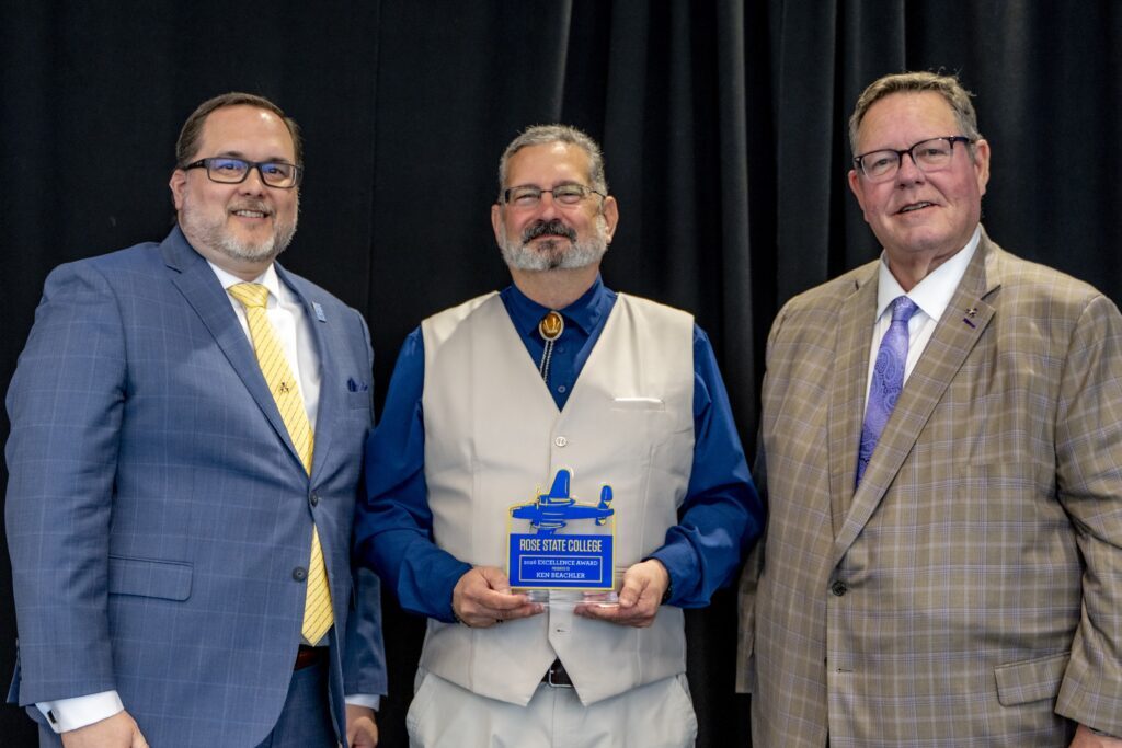 Three men pose together at a Rose State College awards ceremony, with the man in the center holding a 2026 Excellence Award plaque.