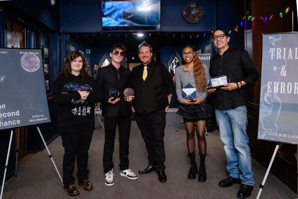 Four student filmmakers and faculty member pose with awards during the Rose State College Film Capstone Awards event, with movie posters displayed on easels in the background.