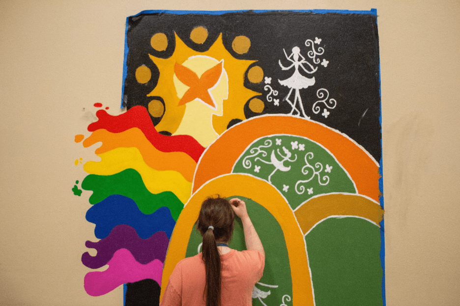 Student paints a colorful mural featuring rainbow waves, abstract figures, and decorative patterns on a wall during an art project.