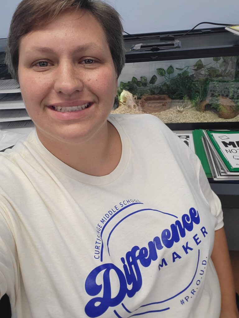 Smiling educator wearing a white “Difference Maker” T-shirt takes a selfie in a classroom or office setting, with a terrarium and educational materials visible in the background.