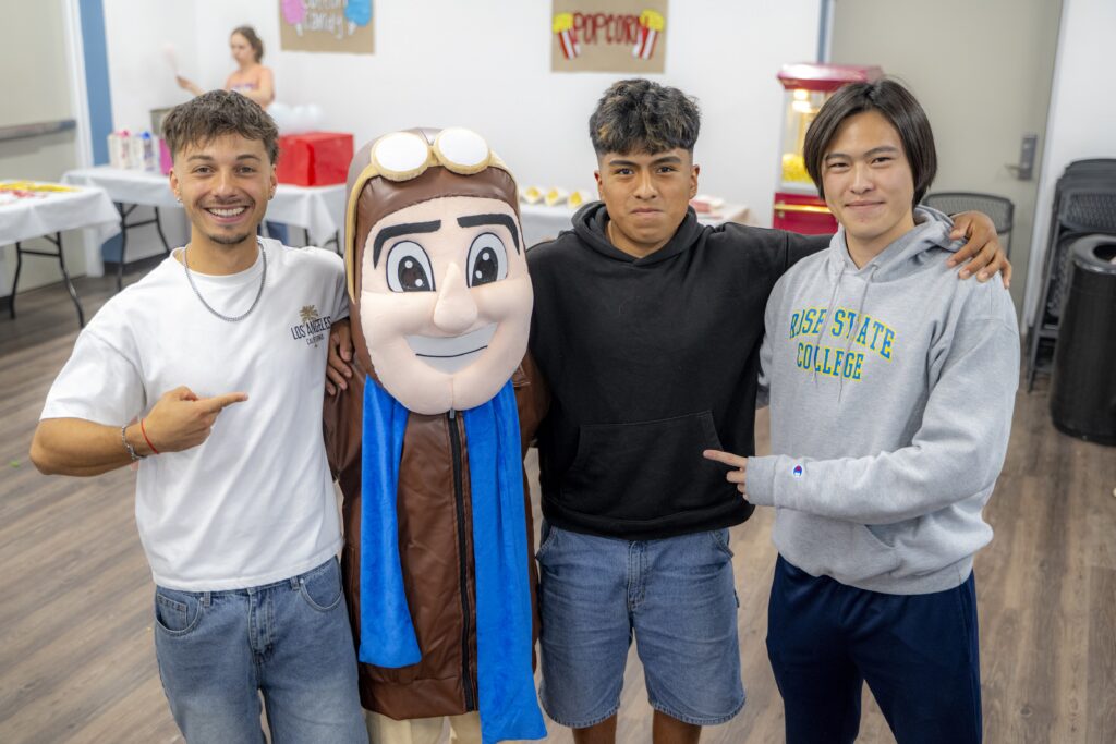 Three students pose with the Rose State College Raider mascot during an indoor residence hall carnival, smiling with their arms around each other as event activities and refreshments are visible in the background.