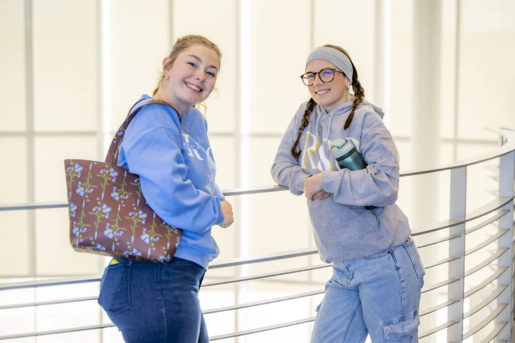 Two students stand and chat along an indoor balcony railing, smiling toward the camera while carrying personal items between classes.