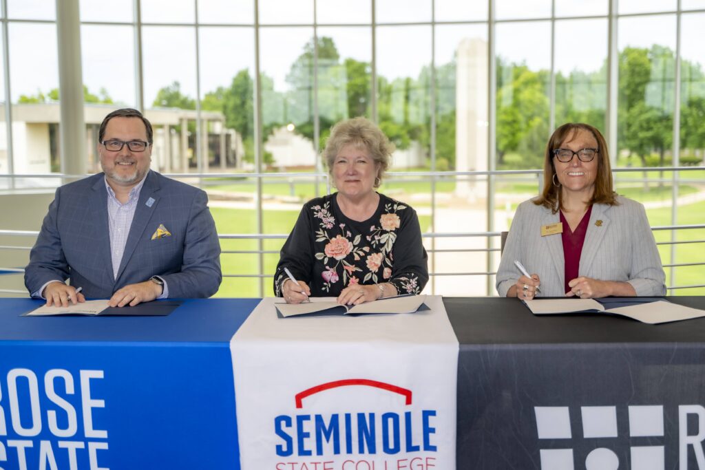 **Alt text:** Three representatives from Rose State College and Seminole State College sit at a table signing partnership documents during a memorandum of understanding ceremony, with institutional banners displayed in front of them and large windows overlooking campus in the background.