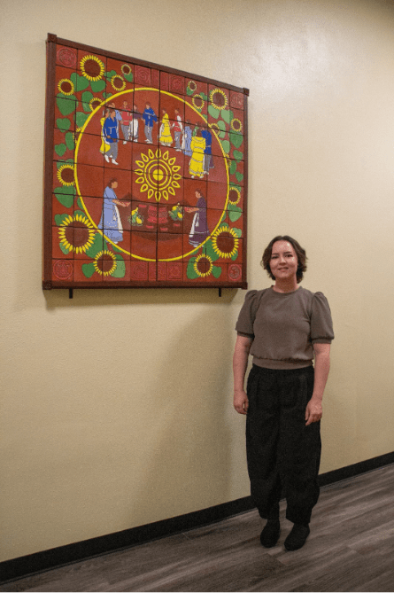 A person stands beside a large colorful wall artwork featuring sunflowers, community figures, and a central sun motif displayed in a campus building hallway.