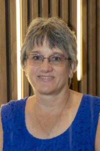 Professional headshot of a smiling woman with short gray hair and glasses wearing a royal blue sleeveless top against a wood-paneled background with vertical lights.