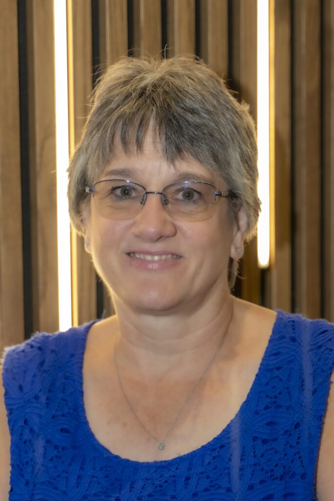 Professional headshot of a smiling woman with short gray hair and glasses wearing a royal blue sleeveless top against a wood-paneled background with vertical lights.