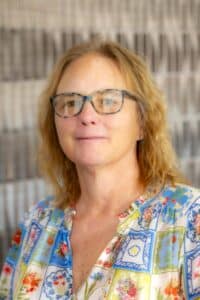Professional headshot of a woman with shoulder-length blonde hair and glasses, wearing a colorful floral blouse, in front of a softly blurred neutral background.