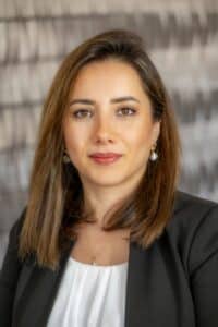 Professional headshot of a woman with straight brown hair, wearing a black blazer and white blouse, in front of a softly blurred neutral background.