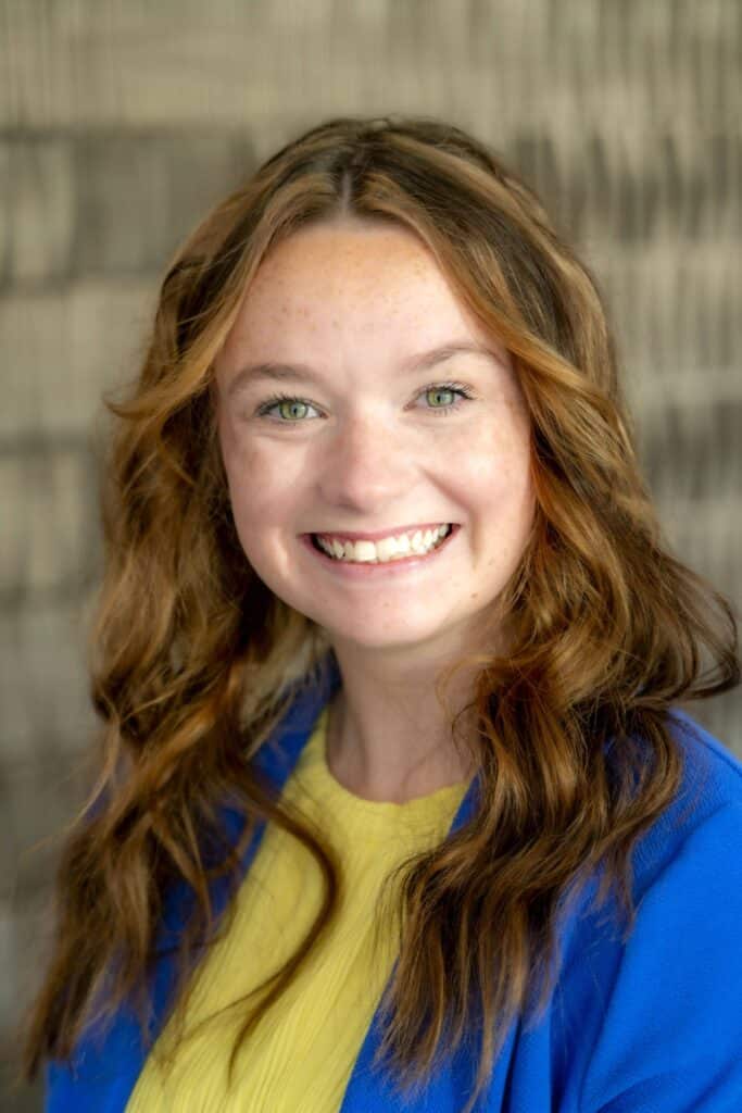 Professional headshot of a smiling woman with wavy light brown hair, wearing a yellow top and royal blue blazer, in front of a softly blurred neutral background.