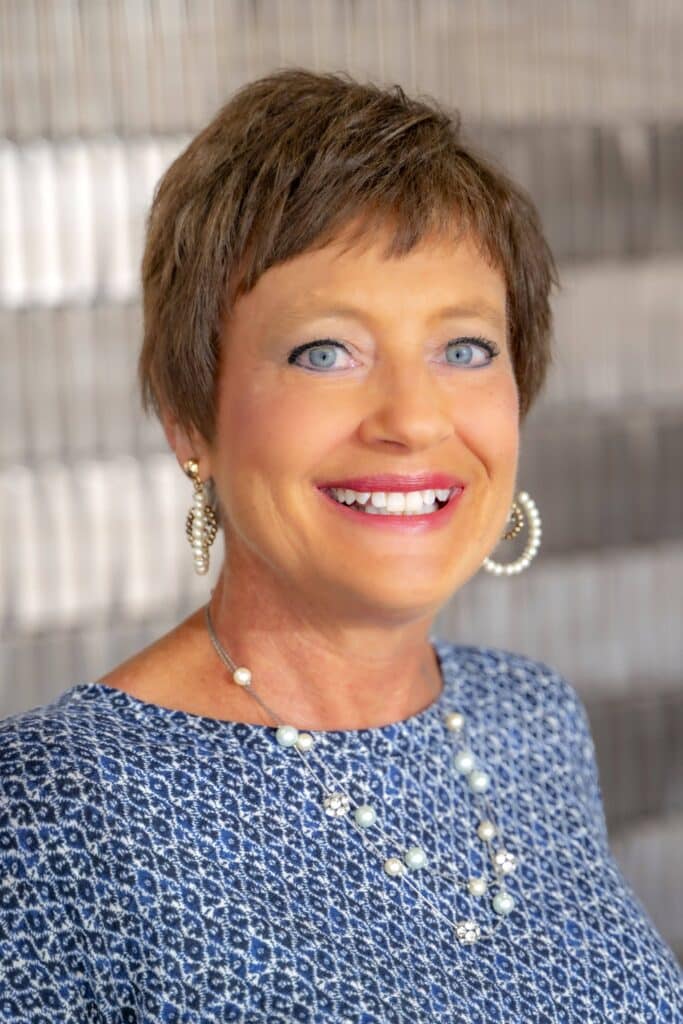 Professional headshot of a smiling woman with short brown hair, wearing pearl jewelry and a blue patterned blouse, in front of a softly blurred neutral background.
