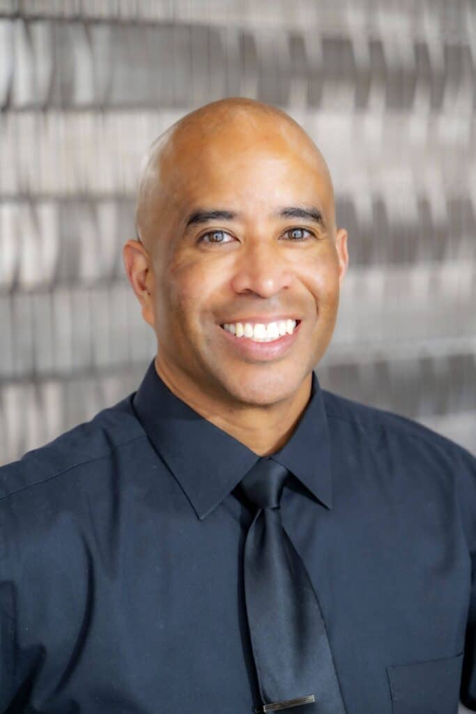 Professional headshot of a smiling man wearing a black dress shirt and black tie, in front of a softly blurred neutral background.