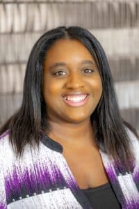Professional headshot of a smiling woman with straight dark hair, wearing a white and purple patterned blouse, in front of a softly blurred neutral background.