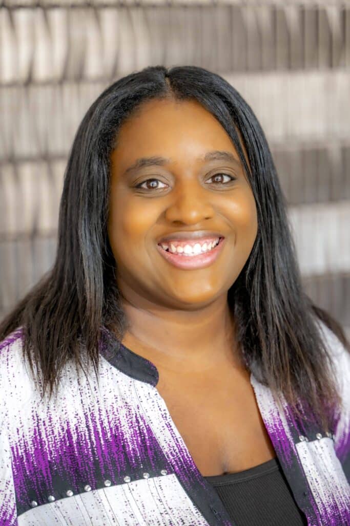 Professional headshot of a smiling woman with straight dark hair, wearing a white and purple patterned blouse, in front of a softly blurred neutral background.