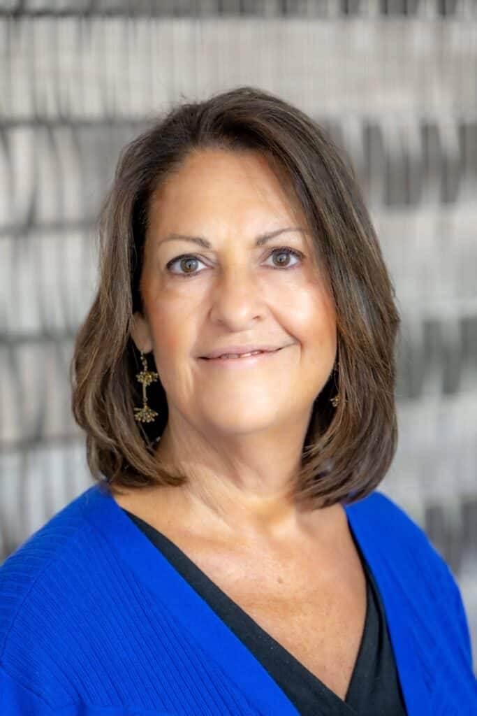 Professional headshot of a woman with short brown hair wearing a royal blue cardigan and black top, in front of a softly blurred neutral background.