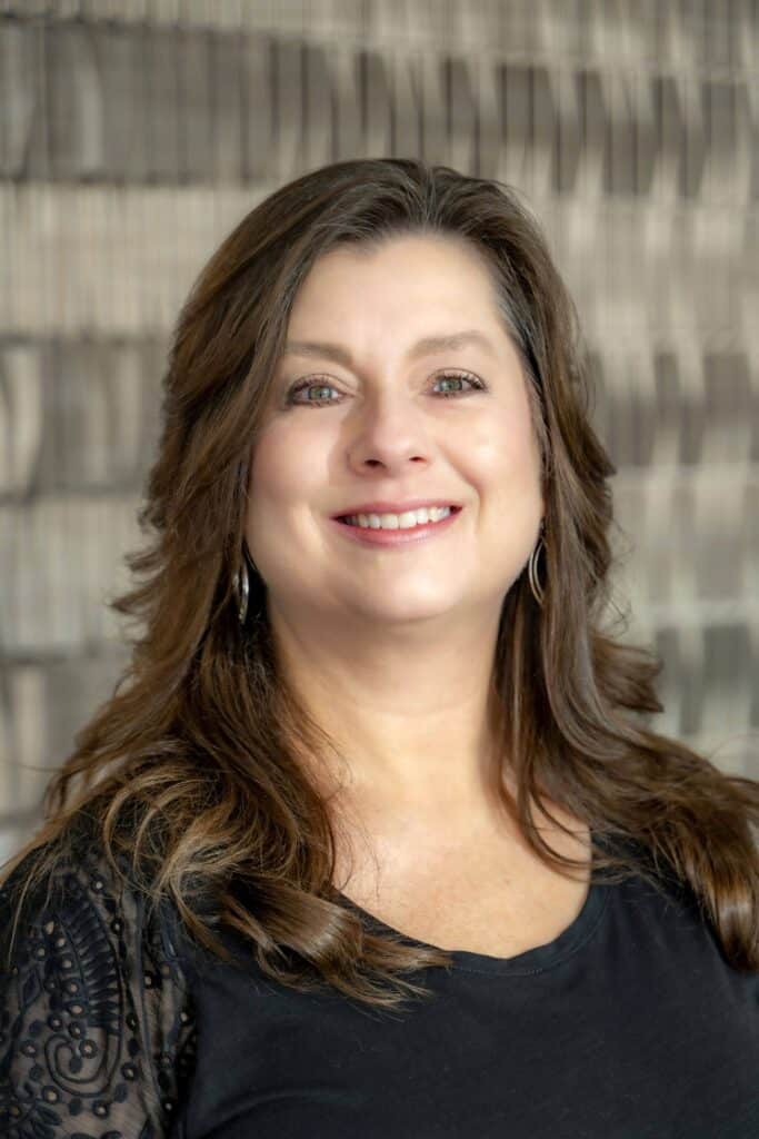 Professional headshot of a smiling woman with long brown hair wearing a black blouse with lace sleeves, in front of a softly blurred neutral background.