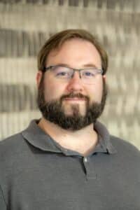 Professional headshot of a man with short brown hair, glasses, and a beard wearing a dark gray collared shirt, in front of a softly blurred neutral background.