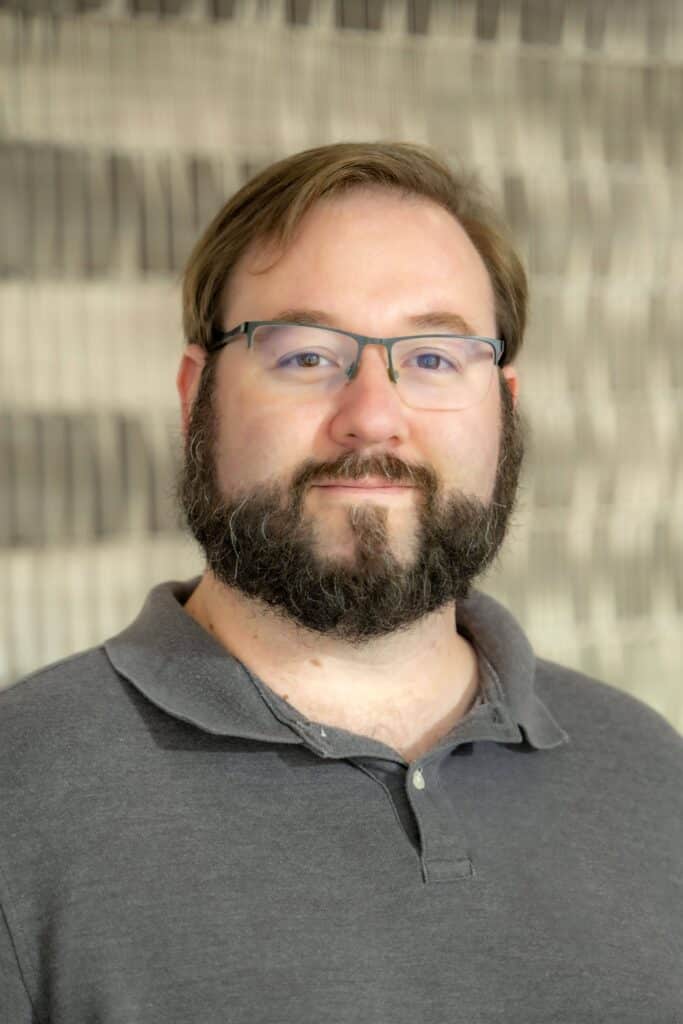 Professional headshot of a man with short brown hair, glasses, and a beard wearing a dark gray collared shirt, in front of a softly blurred neutral background.