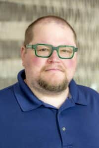 Professional headshot of a man wearing green glasses and a navy blue collared shirt, in front of a softly blurred neutral background.