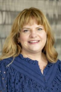 Professional headshot of a smiling woman with blonde hair wearing a navy blue blouse with eyelet details, in front of a softly blurred neutral background.