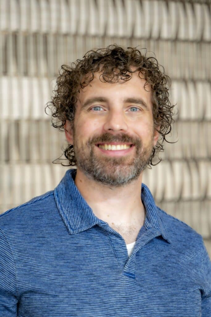 Professional headshot of a smiling man with curly hair and a beard wearing a blue collared shirt, in front of a softly blurred neutral background.