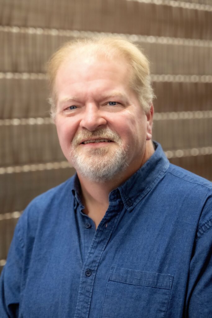 Portrait of a man with short light hair and a beard wearing a blue button-up shirt, smiling in front of a blurred brick wall background.