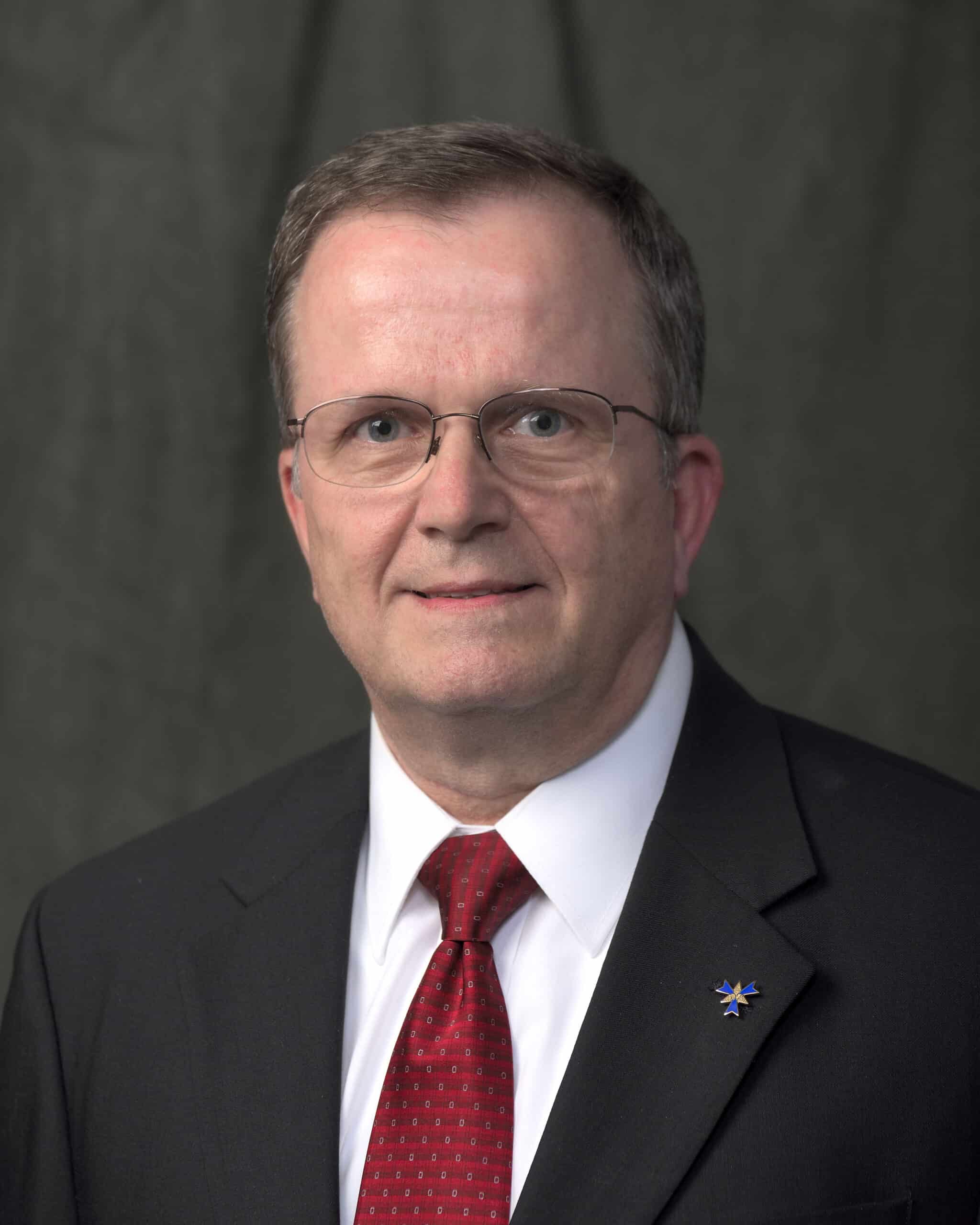 Professional headshot of a man wearing glasses, a dark suit jacket, white dress shirt, and red tie, posed in front of a plain dark background.