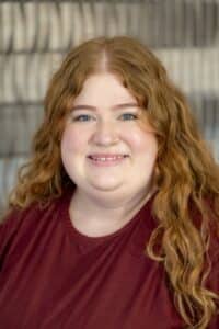 Professional headshot of a smiling woman with long wavy red hair, wearing a maroon top, in front of a softly blurred neutral background.