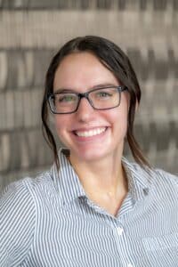 Professional headshot of a smiling woman with dark hair pulled back, wearing glasses and a blue-and-white striped button-up shirt, in front of a softly blurred neutral background.