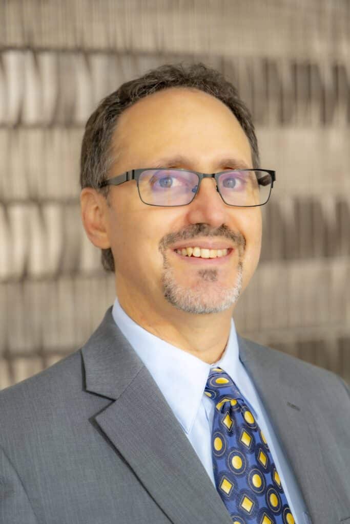 Professional headshot of a man with glasses and a beard, wearing a blue Rose State College polo shirt, in front of a softly blurred neutral background.