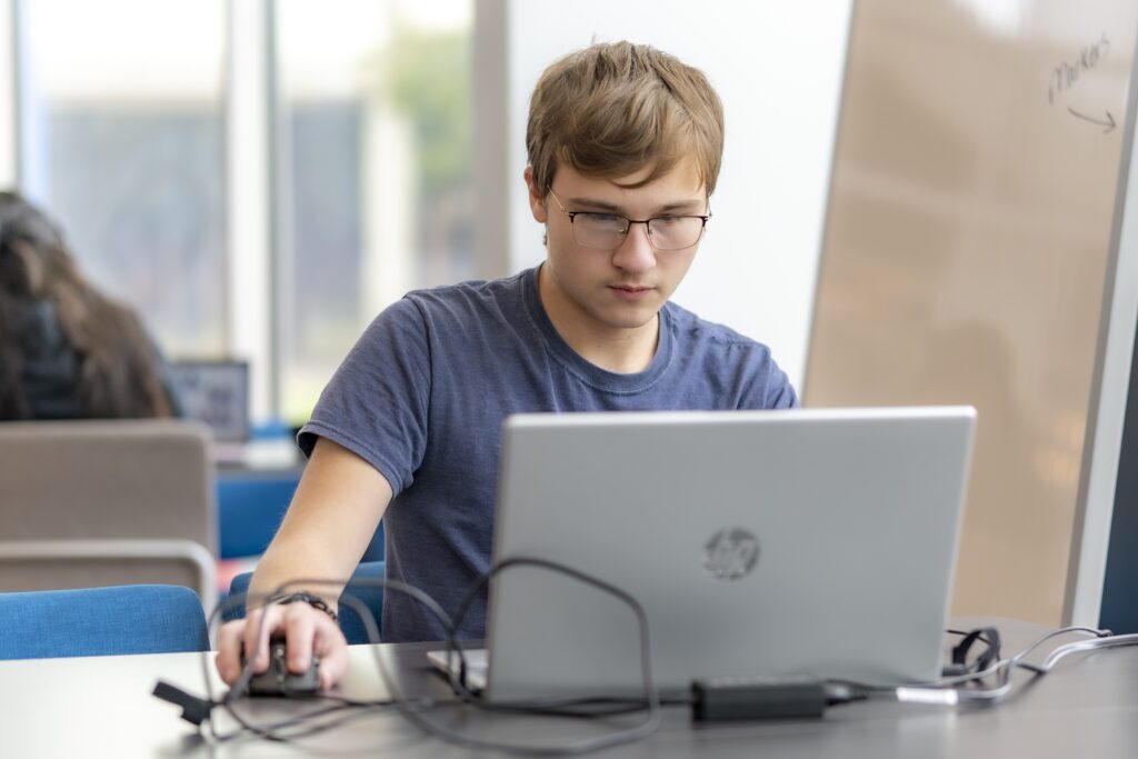 Student works on a laptop at a table in a campus study area, using a computer mouse and focusing on the screen.