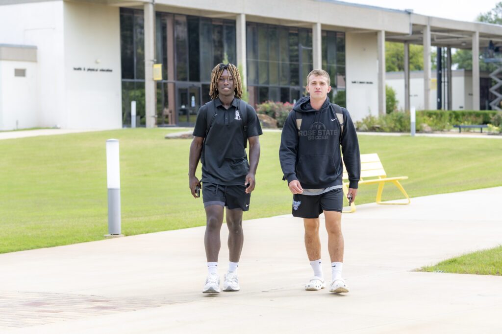 Two Rose State College students walk along a campus sidewalk in front of a building, wearing backpacks and athletic clothing.