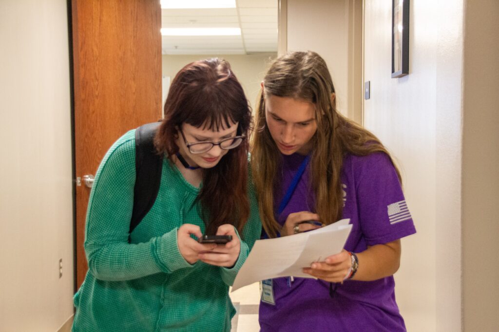 Two Rose State College students look at a phone and a printed paper while finding their way in a campus hallway.