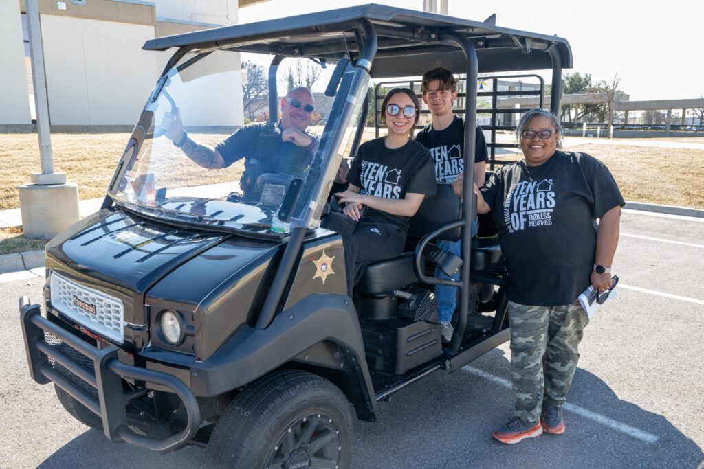 Campus police officer and three students sit and stand around a utility vehicle on campus, smiling at the camera.