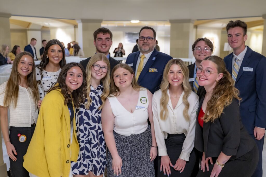 Group of students and a staff member pose together at a college event, smiling and wearing business or business-casual attire with name badges.
