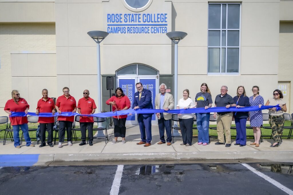 Group of campus leaders, staff, and community members hold a blue ribbon during a ribbon-cutting ceremony outside the Rose State College Campus Resource Center.